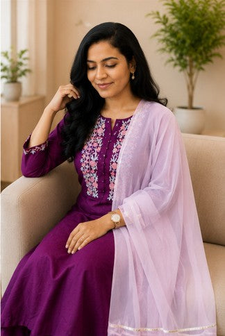 Woman in a purple dress with floral patterns and a light pink dupatta sitting on a beige couch.